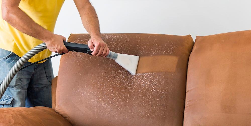 A person uses a vacuum to clean a brown fabric couch.