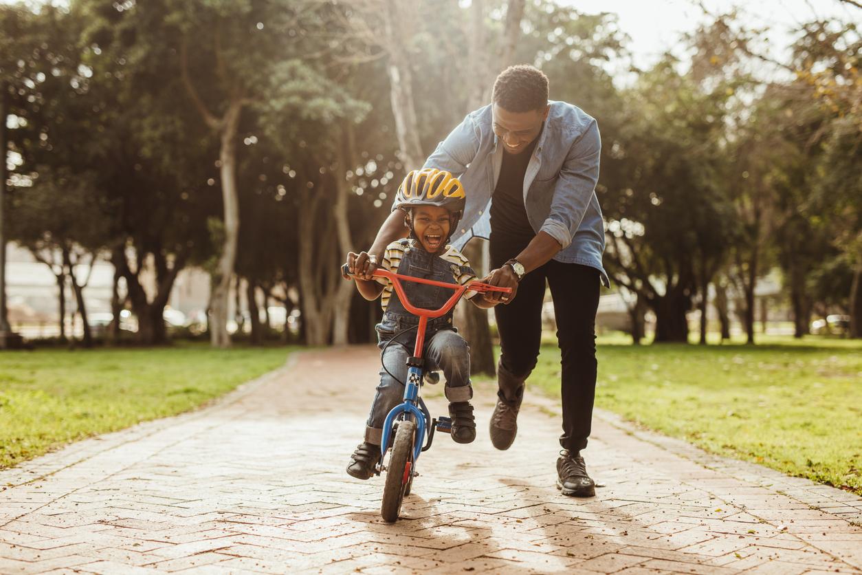 A smiling father steadies his smiling young son on a bicycle in the park.